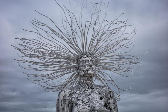 Auto-generated description: A sculpture depicting a human figure with an array of metal wires extending outward from its head against a cloudy sky.