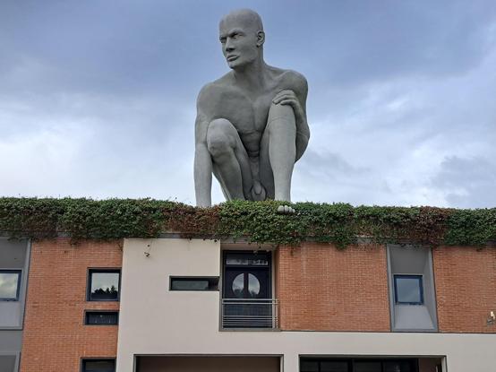 Sul tetto di un edificio ricoperto da siepe si staglia la statua di un uomo calvo, accucciato, nudo.
