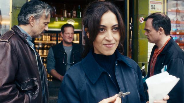 Mehrere Menschen stehen vor einer Trinkhalle Laden mit Regalen voller Waren im Hintergrund. Eine Person im Vordergrund trägt einen dunkelblauen Mantel und hält einen Schlüssel in der Hand. Eine andere Person links trägt eine braune Lederjacke, eine weitere Person rechts einen schwarzen Mantel und hält weiße Papiere oder eine Mappe. Im Hintergrund ist in der Trinkhalle ein Mann in dunkler Kleidung zu sehen. (Bild: HR/Bluelaserboys/Triple Pictures)