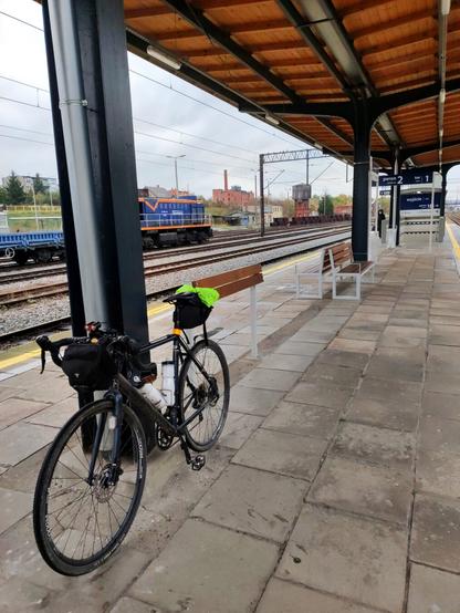 O słup na peronie dworca kolejowego oparty jest ciemnoniebieski rower szutrowy. Za kierownicy i pod siodłem ma czarne torby. Na wierzchu tylnej przyczepiona jest jaskrawożółta kurtka przeciwwiatrowa. W tle widoczna niebiesko-pomarańczowa lokomotywa z wagonami towarowymi.