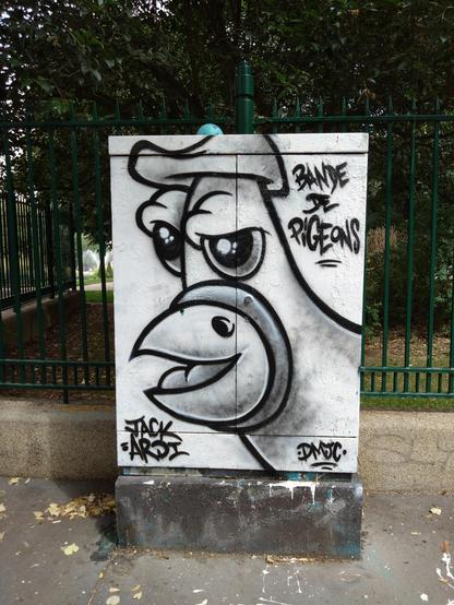 A close up of a pigeon face on an electrical cabinet in front of a garden. It's wearing a hat. The caption reads "Bande de Pigeons".