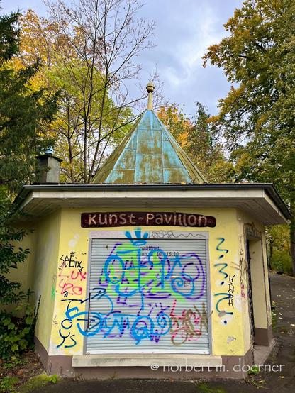 Kleines, gelbes Gebäude mit einem eckigen Kupferdach in einem Park umgeben von Bäumen. Über dem geschlossenen grauen großen Rolladen ein Holzschild mit dem Text "Kunst-Pavillon", und alles bedeckt mit mehrfarbigen ungelenken Grafitti-Tags