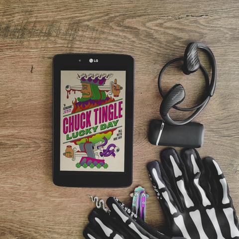 chuck tingle book called lucky day, words at an angle on a stylized and edited king level card, with mirrored images of the king portraying differet gruesome endings they met. black framed lg tablet is placed on a table, with a few items strewn around, such as gloves, lighter, earphones. image and review by wannabekingpin aka vakaris, the reading nosferatu