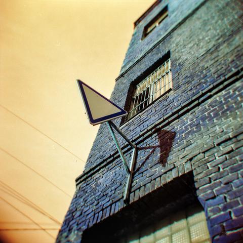 The image shows an upward view of a brick building, captured from a low angle. A triangular road sign with a white center and a blue border is attached to the wall, casting a shadow on the bricks. The building’s windows have metal bars, adding an industrial feel. The sky has a surreal warm orange tone, while the bricks appear in shades of turquoise and blue — a striking color shift typical for Lomochrome Turquoise film. The dreamy tones and slight vignette give the photograph a distinct, almost otherworldly atmosphere, enhanced by the soft focus and unique optical quirks of the Holga camera.
