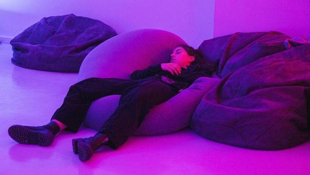 Image: installation shot; photo by Katie O’Neill | we see a figure, apparently asleep, lying on a large bean bag in what may be a gallery space; the lighting is mauve; the face and long hair of the figure are more or less centred horizontally in the photo, but about one third from the top; the head is turned a bit to their right, the right hand is just below the chin and we see the right arm, clad in something dark – all the clothese are dark, including the jeans on the legs; black boots stretch towards us and to the left, soles exposed • “Libraries of Rest by Ciara Barker is an immersive exhibition that invites visitors to imagine the future of restful spaces and practices.    Libraries of Rest debuted at transmediale studio in Berlin as part of Zeitgeist Irland 24, where audiences were encouraged to consider speculative futures centred on collective well-being, divorced from productivity. Facilitating the creation of a shared resource of ideas for accessing rest, the second…”