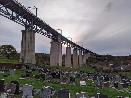 Im Vordergrund eine nach rechts leicht abfallende Wiese, auf der in vier bis fünf Reihen Grabsteine stehen. Dahinter die oberen zwei Drittel des Bildes einnehmend, mehrere wuchtige, eckige Betonpfeiler, deren oberen Enden durch eine blau und graue Metallkonstruktion verbunden sind. Auf der Metallkonstruktion Stromleitungen zwischen torähnlichen Metallbügeln. Hinter der Eisenbahnbrücke eine relativ homogene Wolkenfläche, in der Mitte des Bildes schimmert hinter der Brücke leicht die Sonne hindurch.