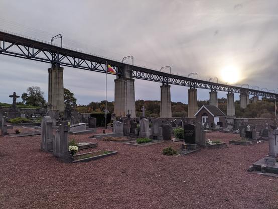 Im Vordergrund eine rote Kiesfläche, auf der zahlreiche Grabsteine stehen. Dahinter die oberen Hälfte des Bildes einnehmend, mehrere wuchtige, eckige Betonpfeiler, deren oberen Enden durch eine Metallkonstruktion verbunden sind. Auf der Metallkonstruktion Stromleitungen zwischen torähnlichen Metallbügeln. Hinter der Eisenbahnbrücke eine relativ homogene Wolkenfläche, rechts im Bild schimmert direkt über der Brücke leicht die Sonne hindurch.
