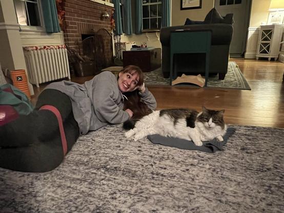 A person lies on their side on a rug, smiling and looking at the camera, while a fluffy cat rests beside them on a small blanket. The setting is a cozy living room with wooden floors and warm lighting.