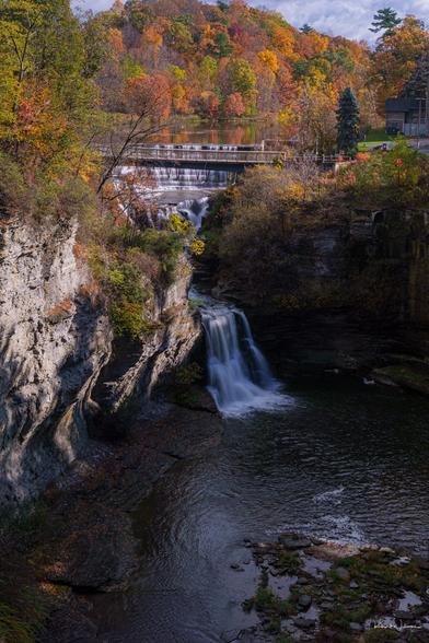 Triphammer Falls cascading in two tiers from Beebe Dam spillway into Fall Creek Gorge, framed by layered sedimentary rock walls and brilliant autumn foliage in oranges and golds, with footbridge visible at top
