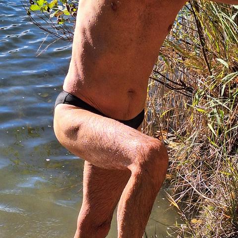 Der bloße Körper eines Mannes mit schwarzer Badehose vom Brustbereich bis zum Knie bzw. Knöchel, der aus dem Wasser steigt. Rechts Uferböschung, links Wasseroberfläche mit leichten Wellen auf denen das Sonnenlicht reflektiert. Die Farbe des Seewassers changiert von graugrün unten zu blaugrau oben.