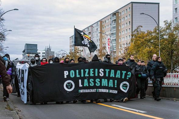Das Foto von © Björn Obmann zeigt die von Bullen eskortierte Demo mit Frot- und Seitentransparenten. Auf dem Fronttransparent steht der Text: "Polizeistaat? Lassma!", darunter "Patriarchat sterben lassen"