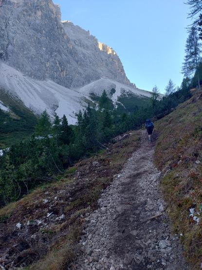 I walk upon a gravel path. There is a wooded section in front me. Far rising Dolomites on the left. A small and upper part of the mountains glows in the sunlight.