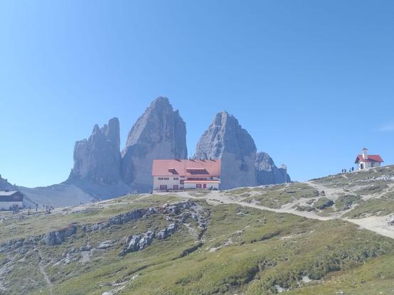The Dreizinnenhütte sits in a vast and open are in front of the Tre Cime di Lavaredo. People can be seen around the hut. The sky is clear blue.