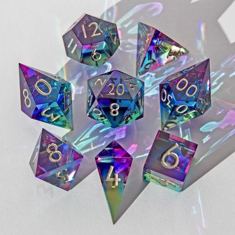 Ein 8-teiliges Würfelset in Blau, Lila und Gelb und Grüntönen. Das Set ist glasklar und durchsichtig und hat dadurch einen Glaseffekt. Die Würfel liegen auf weißem Untergrund und werden durch die Sonne erleuchtet, was zu Reflektionen auf dem Untergrund führt.  Auf dem Bild liegen alle Würfel mit der höchsten Seite nach oben und sind um den D20 herum angeordnet. Das Set besteht aus D4 Shard, D4 Pyramide, D6, D8, D10, D00, D12 und D20. Die Nummern sind in einem Platingold bemalt.