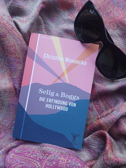 Foto des Buches, auf dessen Cover drei hintereinandergestaffelte stilisierte Hügel in Violett- und Blautönen unter einem rosafarbenen Himmel zu sehen sind, in den so etwas wie drei Lichtstrahlen hineinragen – aus diesen starken Scheinwerfern, die bisweilen bei Großevents an den Himmel leuchten. Das Buch liegt auf einem Tuch, dessen Muster vor allem die Rosétöne des Covers aufnimmt, an der oberen rechten Ecke des Buches liegt zudem eine dunkle Sonnenbrille – wichtiges Hollywood-Accessoire!