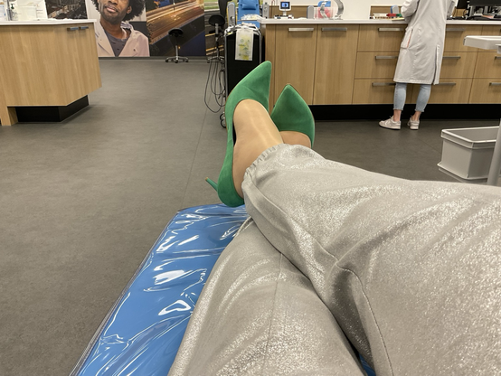 Emma zit ontspannen met haar benen gekruist tijdens het doneren van plasma bij de bloedbank. Ze draagt groene pumps.
