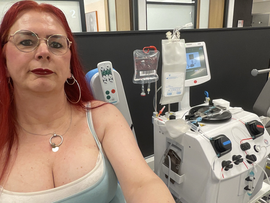Emma, met rood haar en bril, glimlacht ontspannen tijdens haar plasmadonatie bij de bloedbank. Achter haar zijn medische apparaten te zien die gebruikt worden voor het afnemen van plasma. Ze draagt een lichtgroene top.