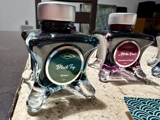 Two bottles of ink sit on a textured surface. The first bottle, labeled "Black Ivy," is a dark teal color with a silver cap, marked as 50ml and categorized as "Sheen." The second bottle, labeled "All the Best” says “shimmer & Sheen”