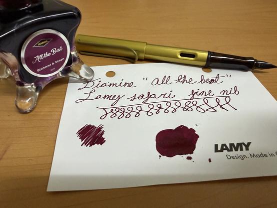 An ink bottle labeled "Diamine All the Best" sits beside a yellow Lamy fountain pen on a wooden surface. A piece of paper displays a handwritten note and ink swatches in a dark burgundy color.