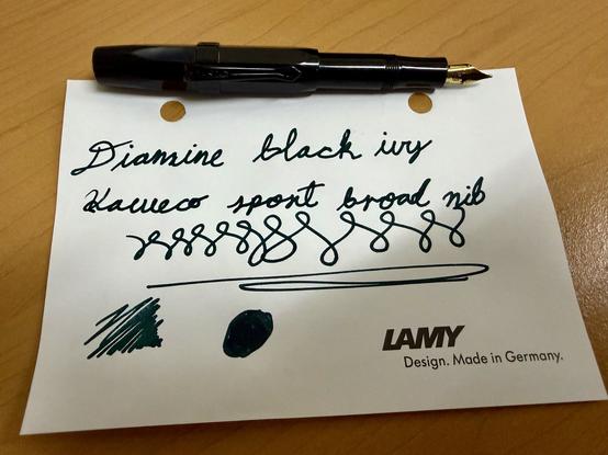 A black fountain pen rests atop a white sheet of paper with handwritten text. The text reads "Diamine black ivy" and “Kaweco sport broad nib." Beneath are various ink swatches, including lines and blobs in green-black.
