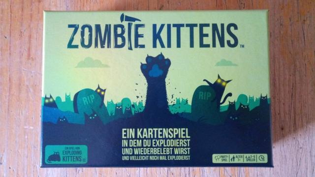 Auf einem Holztisch liegt das Spiel "Zombie Kittens". Das Cover ist in verschiedenen Grüntönen gestaltet, mit einer Katzenpfoten, die zombiemäßig aus der Erde eines Grabes auf einem Friedhof ragt. Drumherum stehen Grabsteine, dazwischen und darauf sind Katzen mit gelb glühenden Augen zu erkennen.