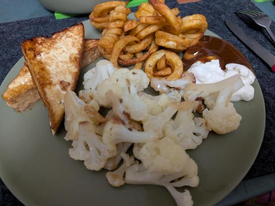 Ein grüner runder Teller, links außen zwei Tofuecken, oben Curlyfries, rechts bzw mittig gebratene Blumenkohlröschen. Oben rechts am Rand Mayo & Curryketchup