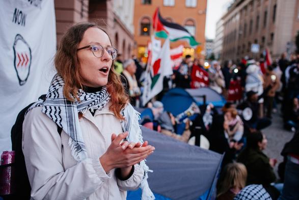 In the front a protester is clapping and raising slogans and in the behind we can see many protesters and some put up tents.