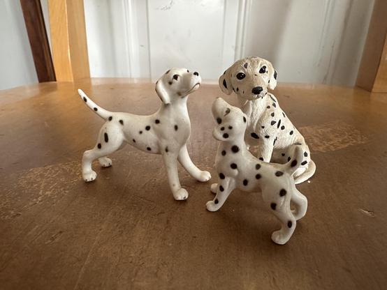 Three small ceramic statues of black spotted dogs, what looks like a sitting dad, an attentive mom, and a wee pup. They are standing on a wood surface and are a bit yellowed with age.