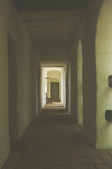 A narrow hallway with white plastered walls, stone floor, and wooden ceiling. At the end of the corridor, soft daylight enters through a doorway, illuminating a single wooden bench placed along the wall. The image evokes quietness and timelessness.