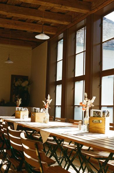 A cozy indoor seating area in a traditional Austrian house. Sunlight streams through tall wooden windows, lighting up wooden tables and chairs arranged neatly in rows. Small vases with dried flowers and wooden boxes with postcards decorate the tables, creating a warm and nostalgic mood.