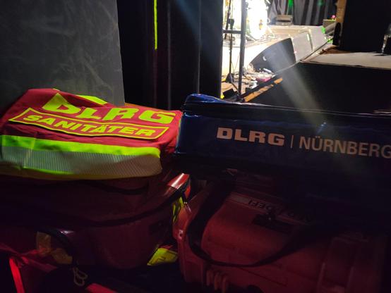 Ein paar Taschen mit sanitätsmaterial auf einer Ablage links liegt eine DLRG Jacke mit Sanitäter Aufschrift.
Dahinter ist eine bühne zu sehen.