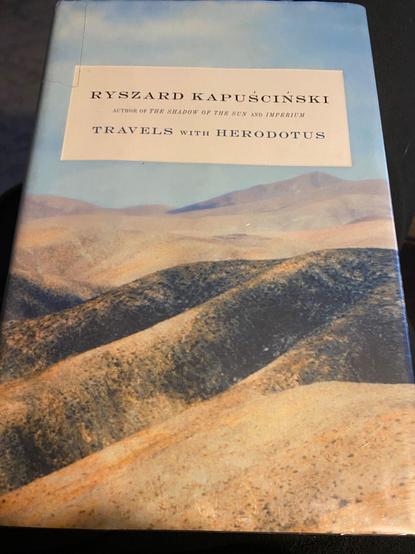 Photo is of a book titled 'Travels with Herodotus' by author Ryszard Kapuscinski in black type in a cream colored text box.  Background is a pastel painting of some hills beneath a pale blue sky.