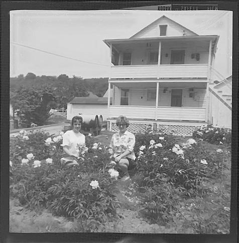 Black and white shot of a backyard. Two women are sitting in a flower bed. There's a two-story house in the background.