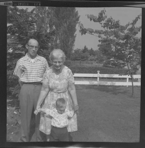 A white-haired woman is holding out the arms of a young boy. Behind her is a rather serious-looking gentleman. Black and white photo.