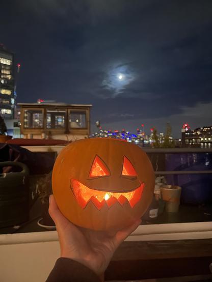 A hand holds a carved, glowing pumpkin with a toothy grin and triangle eyes carved into it. Behind it is a wheelhouse and deck of a Dutch barge, a nighttime city skyline, and a moon shrouded in clouds.