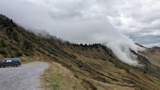 Blick auf eine Bergstraße am Furkapass im Bregenzer Wald. Links steht ein dunkler Van auf einem Schotterparkplatz, rechts ziehen Nebelschwaden über die grünbraunen Hänge und Nadelwälder. Der Himmel ist grau und wolkenverhangen, die Szene wirkt ruhig und mystisch.