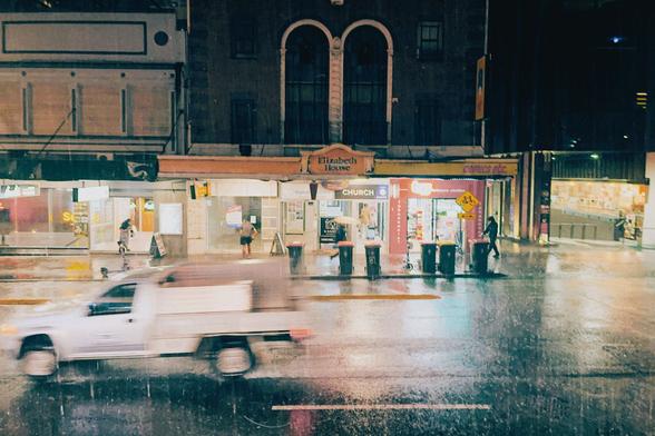 A ute passes by on a rainy night in the city as pedestrians scramble for cover under shops canopies. The rubbish bins stand in the pouring rain unbothered.