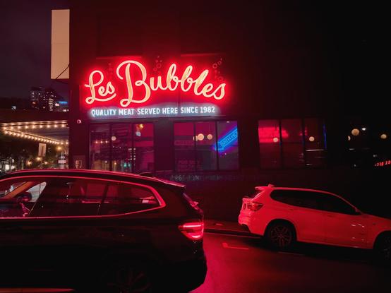 A neon sign says, “Les Bubbles” and declares “Quality meat served since 1982”, lighting the cars with a red glow.