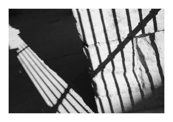 Photo noir et blanc. Ombre portées d'une grille sur un mur en pierre et un sol de terre battue dans les arènes d'Arles. Un triangle d'ombre sépare les 2 jeu de rayures.