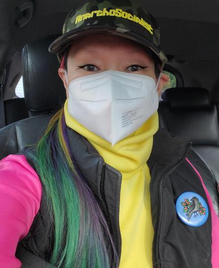 Me, in a car, wearing a white KN95 mask, a yellow turtleneck, a black puffer jacket over a pink top, and a camouflage hat with "AnarchoSocialist" in yellow lettering. My hair is colored green, purple, and yellow. A blue button with Indigenous raven and berries art is pinned to my black puffy vest.