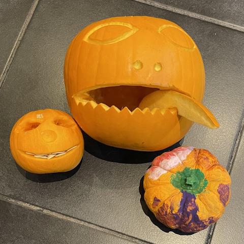 Three orange carved pumpkins indoors on the floor. Small with a face and white seeds as random teeth, medium in the middle with a face and tongue sticking out, and a small painted one on the right.