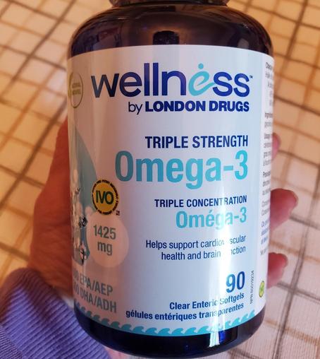 A person's hand holding a dark blue bottle of "wellness by LONDON DRUGS Triple Strength Omega-3" supplements. The label indicates triple concentration, supports cardiovascular and brain health, and contains 90 clear enteric softgels.