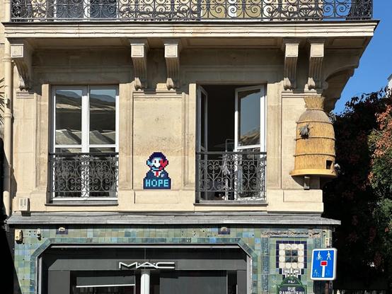 first-floor windows (full height, with wrought-iron French balconies) above a shop. The right one is partially open. Next to it, on the corner of the building, is a sculptural ornament of a beehive. Between the windows is a tiled pixel-art mosaic looking like a homage to the Shepard Fairey Obama Hope poster, in the style of the street artist Invader