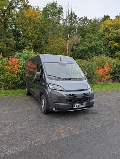 Ein Fiat Ducato in einem blau-grau metallic geparkt vor einer Hecke.