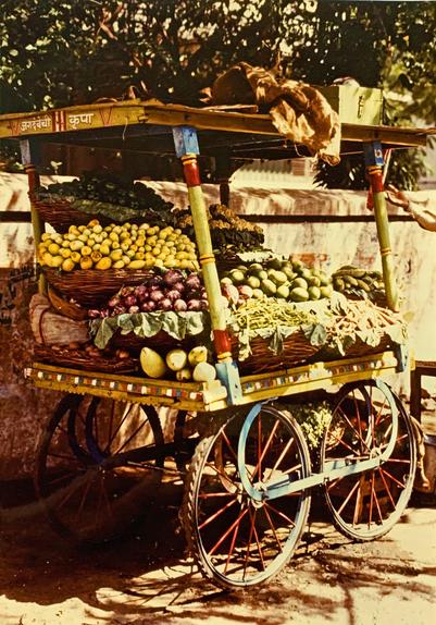 Ein sehr alter Verkaufswagen mit Obst und Gemüse in Indien 1985/86, bunt verziert und liebevoll in Körben aufgeschichtet. Die Räder haben die Größe von Fahrrädern mit roten Speichen. Der Wagen steht an einer Mauer in der Stadt und hat ein Dach.