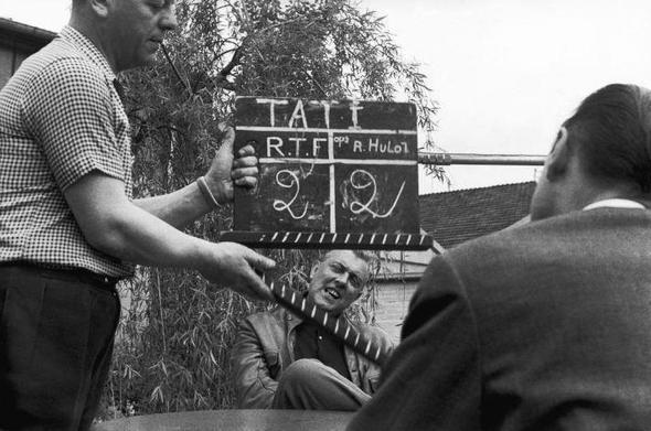 B/W picture of an interview with Tati. Jacques Tati is sitting in the back of the photo (centered). He sits with an interviewer (we see his back). Another man with a clapper is standing, he is about to clap the clapper. We see Tati's head in between the big clapper.