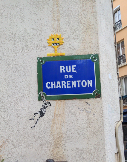 Sur le panneau "Rue de Charenton", une mosaïque représente, en nuances de jaune, un soleil avec des yeux et une bouche, sur un pied avec un socle ; en bas à gauche est collé le dessin en noir et blanc d'une personne, en chemise, short et pieds nus, suspendue en mouvement à la vis du panneau.