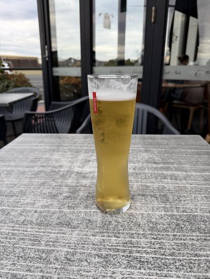 A pint of beer sitting on a table. Not your usual pint glass but taller and thinner, with Peroni written vertically in small white letters on a red background running down from near the top. The beer is not quite full as the owner has already consumed some!