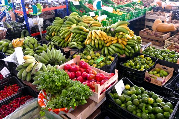 In einer Markthalle. Vor dem Betrachter stehen Kisten mit Bananen, Äpfeln, Limetten, Chilis und vielen anderen Obst- und Gemüsesorten.