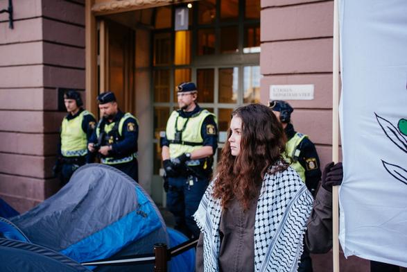 A woman wearing keffiyeh in front of the door and we can see 4 police officers in the background in front of the door. The person is also holding one side of a big banner and between the police and her there are a few blue tents from the protesters.